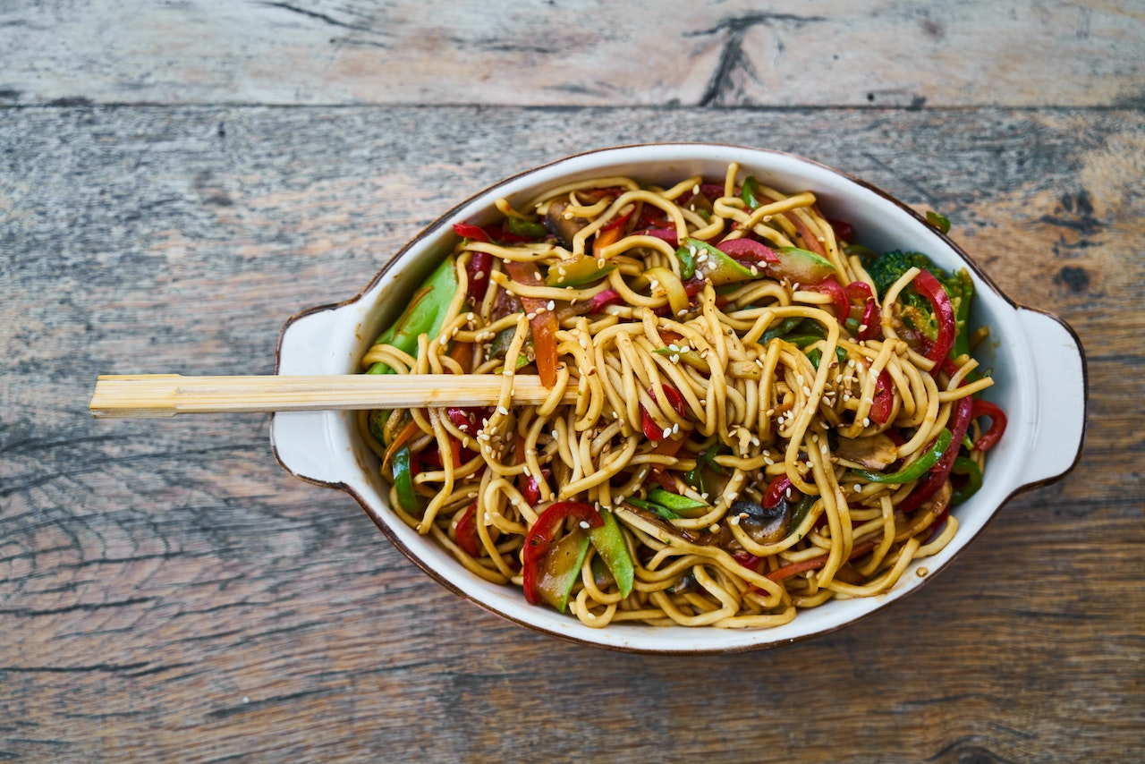 A bowl of noodles and veggies with chop sticks placed on a brown, wooden table